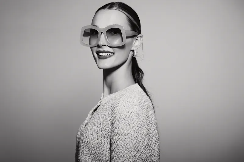 Caucasian woman with fair skin wearing large sunglasses and textured sweater smiles confidently isolated on gray background.