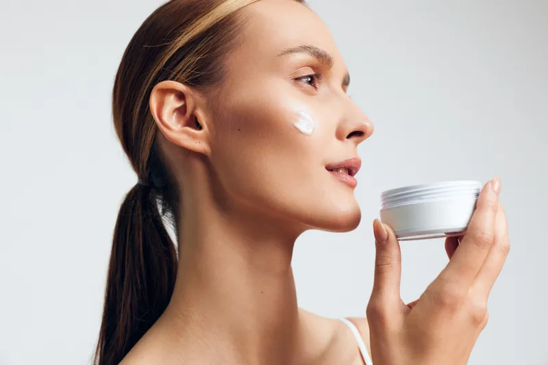 Young woman applies cream from a jar in a close up profile shot, calm mood, skincare routine moment, clean studio backdrop, soft lighting.