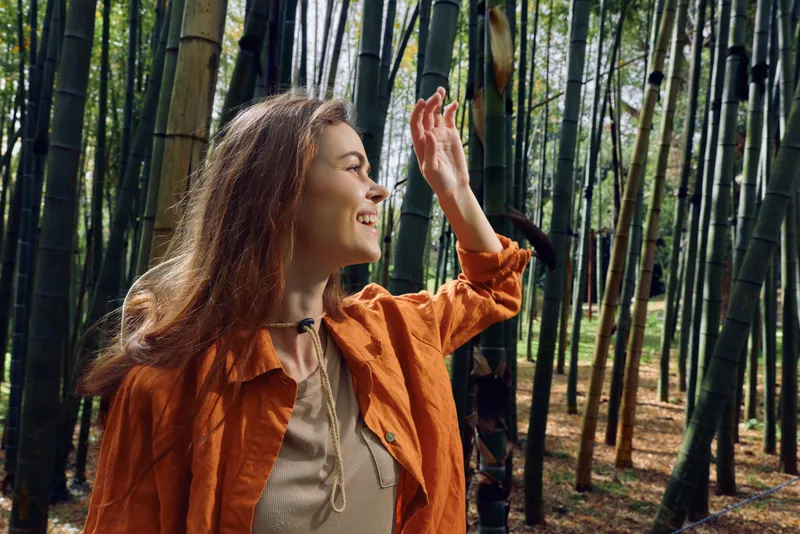 Woman in bamboo forest smiles and reaches up, portrait of a happy traveler in nature wearing an orange jacket, outdoor lifestyle and exploration with sunlight filtering through tall green stalks.
