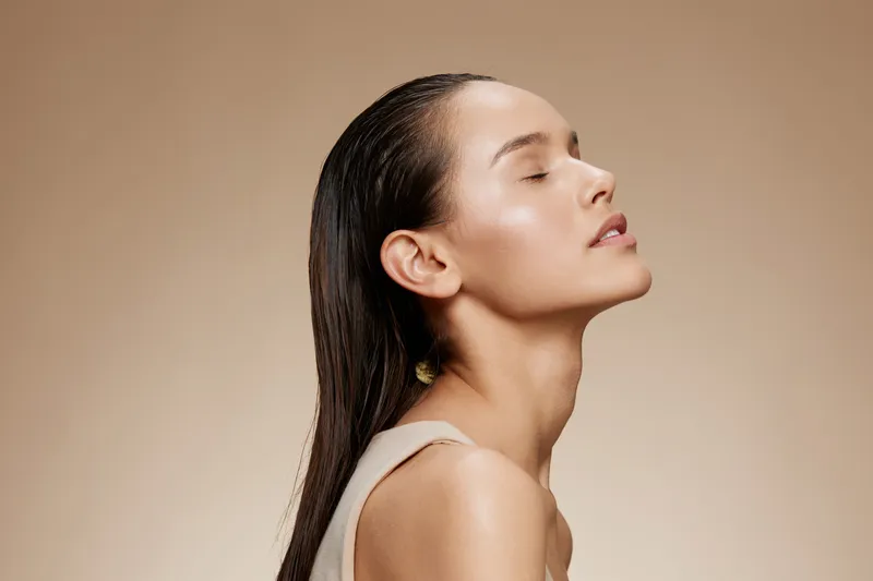 A serene young woman with long, wet hair poses against a neutral background, embodying beauty and tranquility through her peaceful expression and natural skincare routine