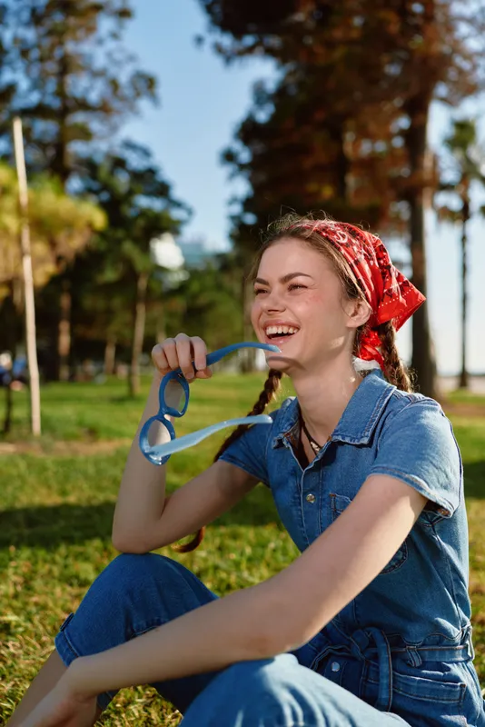 smiling woman holding sunglasses outdoors wearing red bandana and denim outfit sitting on grass in sunny park with trees and blue sky background