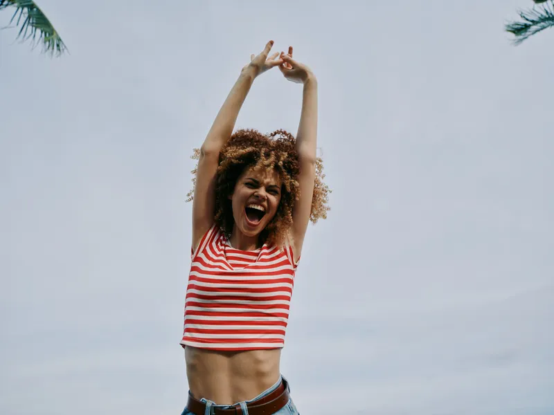 A lively young woman with curly hair raises her arms in celebration against a clear blue sky, wearing a red striped crop top and denim jeans, radiating joy.