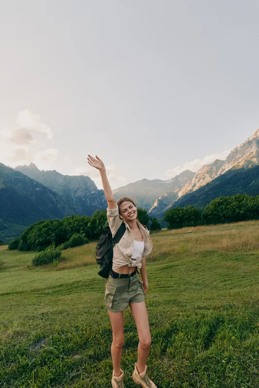 Smiling woman with backpack standing in a grassy field, mountains in the background, ready for outdoor adventure and exploration, sunny and relaxed mood