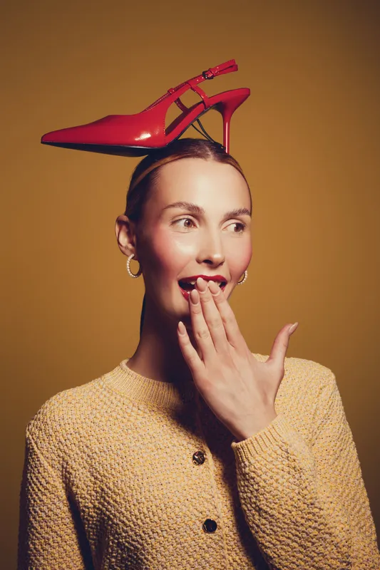 Young woman with red high heel shoe balanced on her head looks surprised with hand near mouth. Studio portrait on yellow background with creative fashion concept and playful mood.