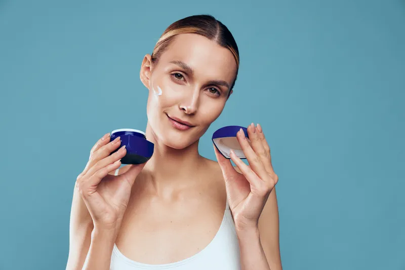 Young woman with clean skin holds open jars of face cream applies moisturizer on cheek with calm expression isolated on blue background beauty skincare concept.