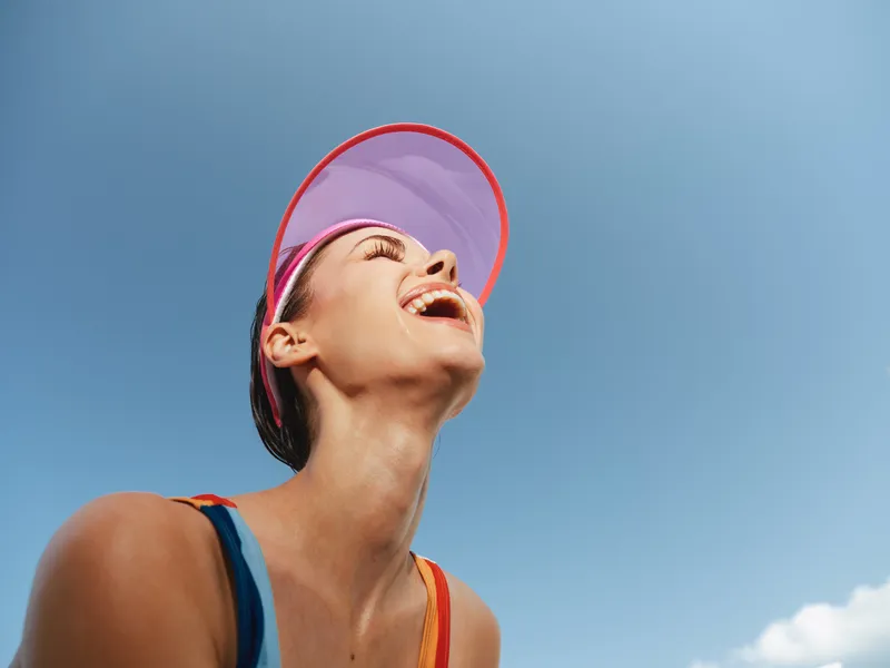 Joyful athlete smiles upward under a pink visor, wearing a bright sporty outfit against a clear blue sky. A vibrant moment of energy, sunshine, and carefree movement.