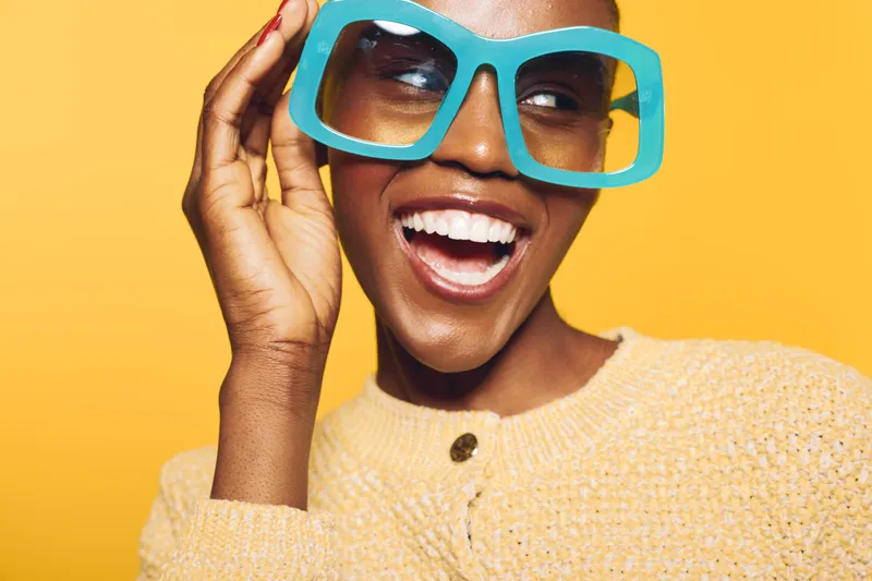 Happy African American woman with dark skin wearing large blue glasses and yellow sweater smiles joyfully on yellow background. Concept for fashion, style, and positive emotions.