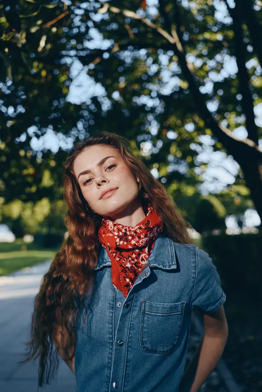 confident woman in denim shirt with red bandana standing outdoors in sunny park with green trees background