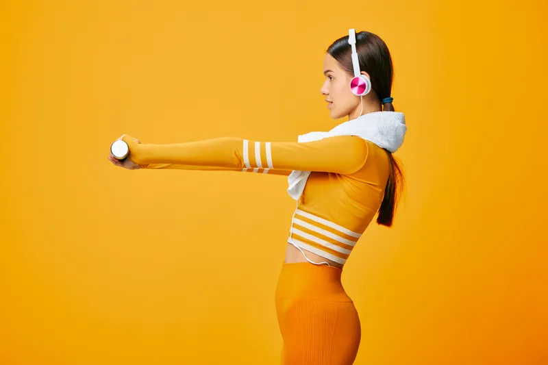 Woman fitness athlete in profile, stretching arms forward with a light weight, wearing orange activewear and headphones against a bold orange studio backdrop.