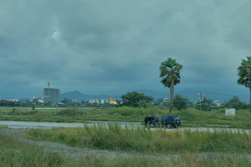 Overcast rural landscape with a lone car on a wet field distant buildings palm trees and hills under a moody sky capturing quiet movement and expansive space in a transportation and nature scene