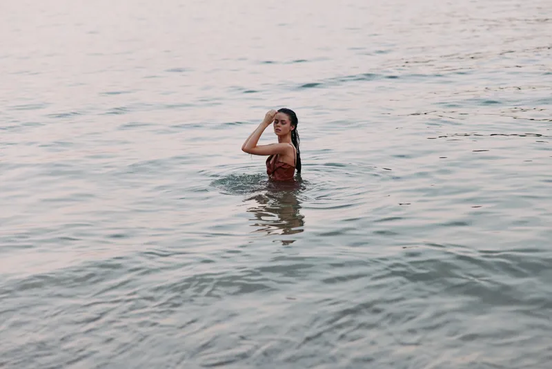A person stands waist deep in calm sea water, turning to look back over shoulder, sunlight glinting on gentle waves, creating a serene summer scene.