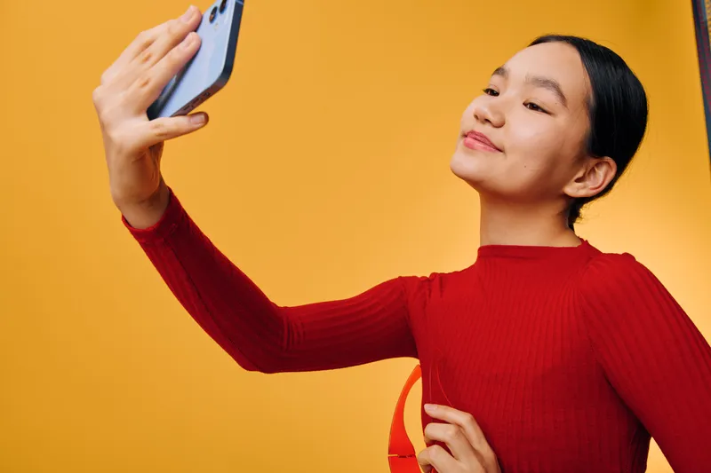 Young woman wearing red sweater taking selfie with smartphone on yellow background. Portrait of happy female holding phone for social media picture with confident expression and modern style.