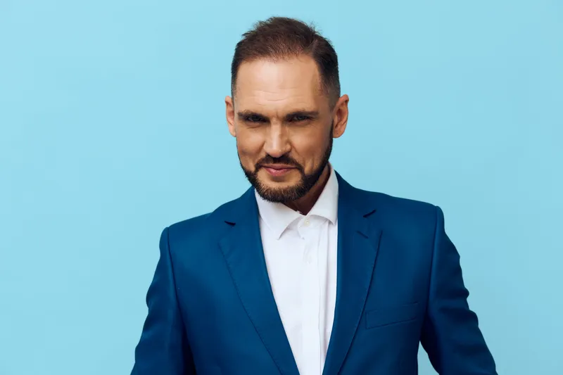 A professional man stands against a solid blue background, a clean studio portrait conveying business poise, focused presence, and ready leadership in a corporate setting.