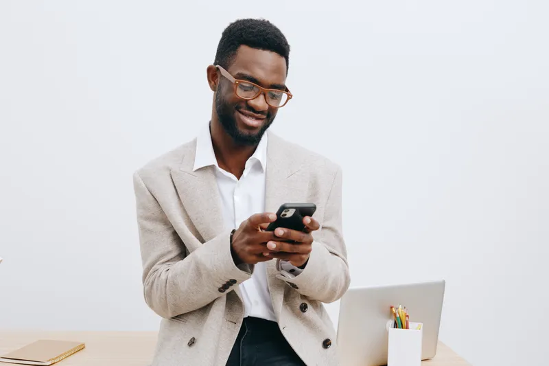 Friendly smiling man with glasses using smartphone in office setting, casual beige blazer white shirt, clean white background, business lifestyle concept