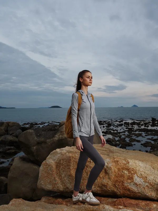 Young woman in sporty attire standing on rocks by the sea, showcasing an active lifestyle with a brown backpack, evoking adventure and exploration