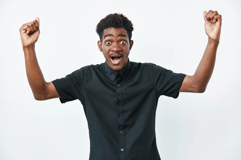 Young black man with dark skin wearing black shirt raising arms and expressing excitement or surprise on white background. Portrait of emotional african male.