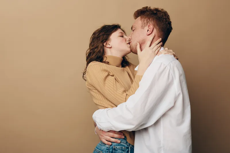 Young couple kissing passionately, embracing each other against beige background. Intimate moment of love, romance, and affection between man and woman in casual clothes.