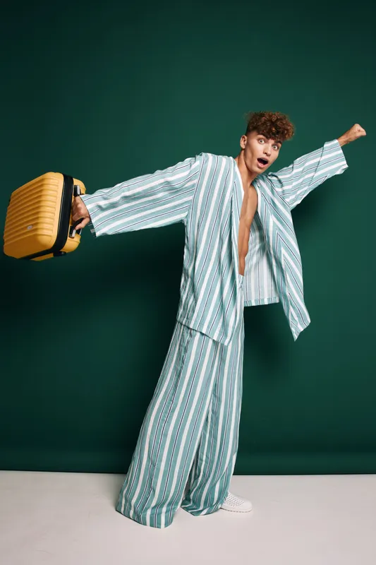 Young man in striped pajamas holding yellow suitcase with excited expression, standing against green background for travel, vacation, and leisure concept.