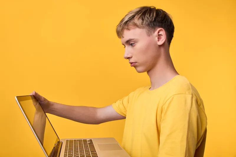 Young male user in a yellow shirt operates a laptop, focused on the screen, against a bright yellow backdrop, conveying remote work, online learning, and technology use in a modern setup for study,