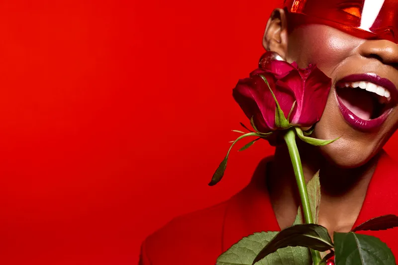 Happy African American woman with dark skin wearing red sunglasses and red jacket holds red rose near face smiling with open mouth on red background. Concept of joy and beauty.