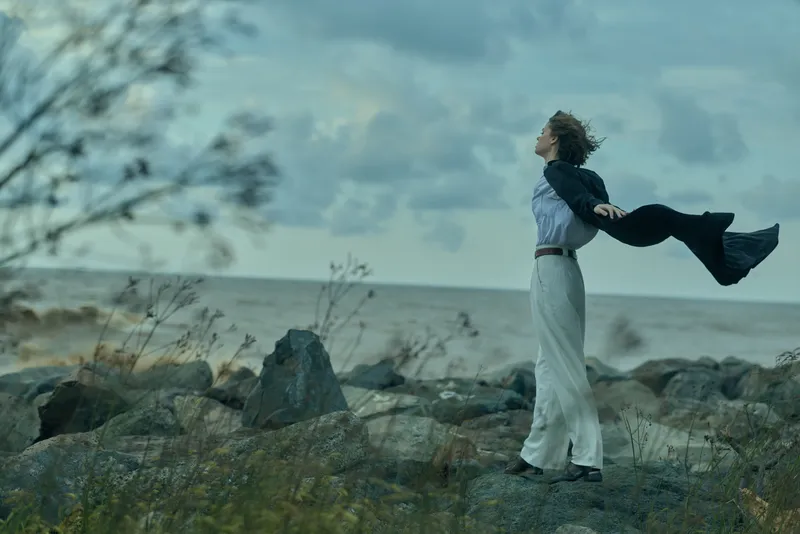 Woman stands on rocky shore with arms outstretched, wind blowing her coat, sea in the background and expansive sky, conveying freedom, resilience and connection with nature