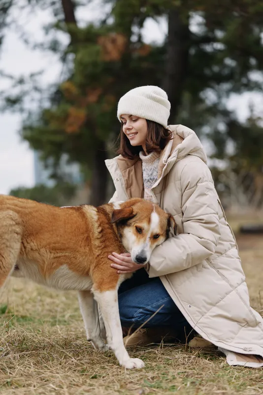 Woman in a puffy coat and knit hat kneels beside a dog in a park, offering gentle touch and warmth on a cool day, with autumn trees visible in the background.