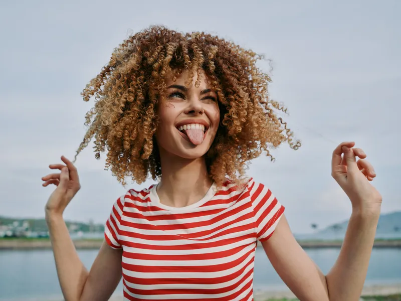 Young woman with voluminous curly hair wears a red and white striped shirt, smiles playfully, sticks out her tongue, and radiates carefree energy beside a sunny lake.