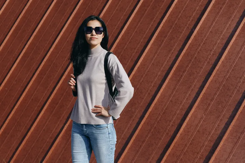 Woman in sunglasses wearing a gray sweater and jeans stands with a backpack against a diagonal wooden wall, exuding casual urban style and confidence, ready for travel, exploration or daily street