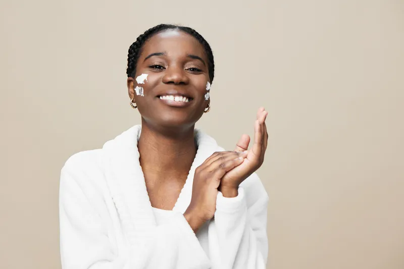 Smiling Black woman applying skincare with cream on her face, wearing a white bathrobe against a neutral beige background