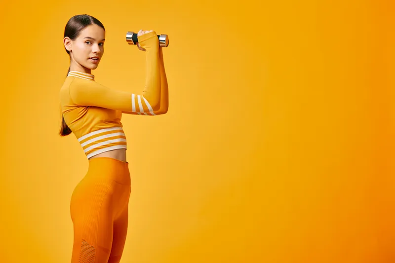 Woman fitness athlete in vibrant orange activewear lifting a dumbbell in a confident profile pose, highlighting toned arms, strong posture and sleek workout clothing on orange backdrop.