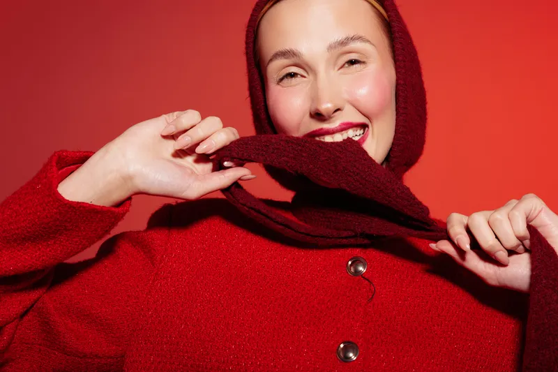 Smiling woman in red sweater and hood poses against red background. Close-up portrait with natural makeup and joyful expression. Concept of fashion, winter style, and happiness.
