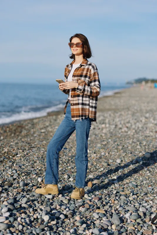 Beach scene with a stylish woman standing on a pebble shore, wearing a plaid shirt, jeans and boots, sunglasses, holding a phone, calm blue sea in the background, relaxed outdoor fashion moment.