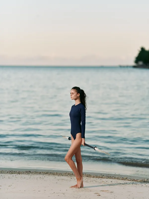 A lone woman stands on a sandy beach in a navy swimsuit, holding a short surfboard beside calm ocean waters as light fades, capturing freedom, balance, and coastal adventure.
