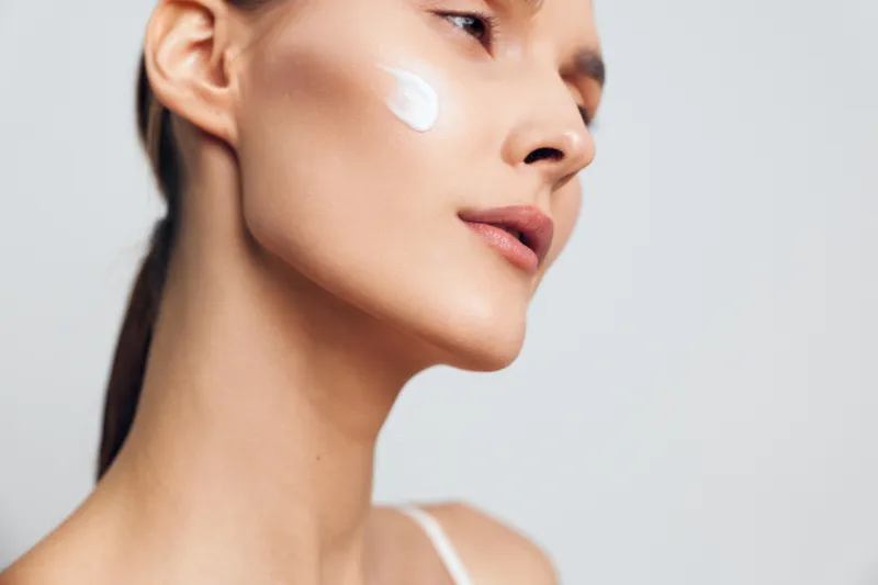 A young woman with clear skin applies white facial cream on her cheek. The close-up beauty portrait shows skincare routine and healthy natural complexion in soft lighting.