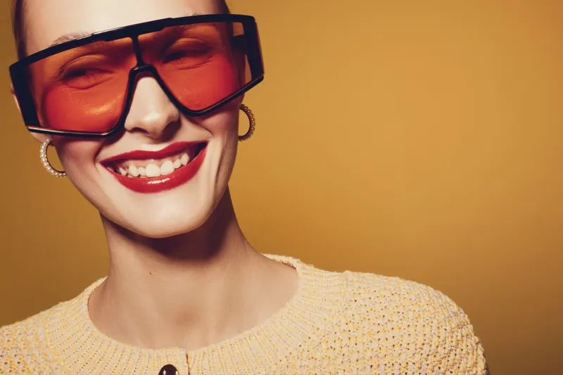 Smiling woman with large orange sunglasses and red lipstick wears yellow knitted sweater on warm brown background. Concept of fashion, style, beauty, and confident female portrait.