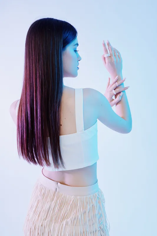 Young woman with long straight hair poses in white top and fringed skirt on light background. Concept of beauty, fashion, elegance, and modern style with soft lighting.