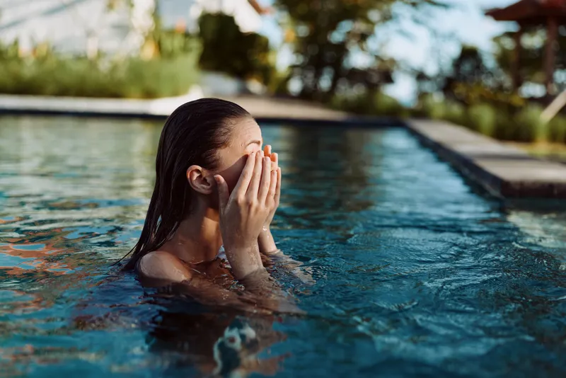 A person with wet hair in a pool covers their face with hands, suggesting stress and relief in a candid lifestyle moment focused on selfcare and calm after a swim.