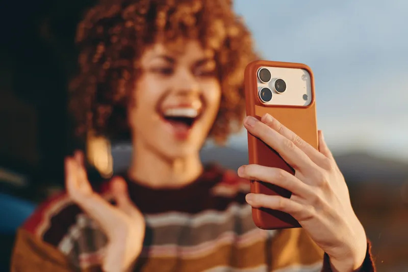 Woman lifestyle, smiling woman with curly hair holds smartphone for selfie outdoors, casual communication and technology, warm orange light, close up portrait, joyful smile