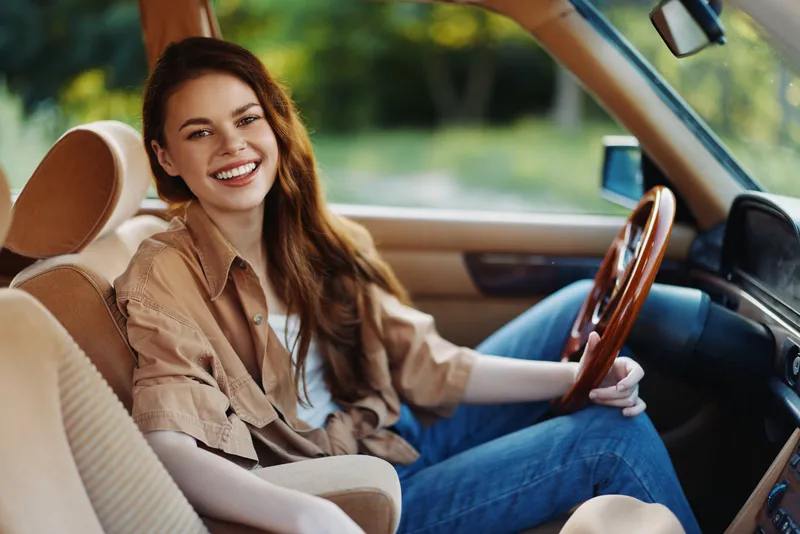 Joyful young woman smiling inside a vintage car, dressed in casual attire with a warm color palette, perfect for lifestyle and travel concepts.