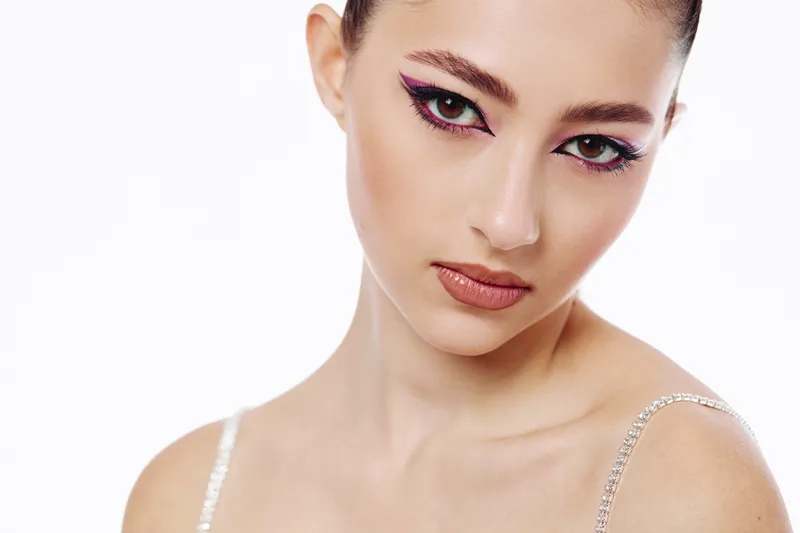 Close-up of a young woman with creative pink eyeliner and glossy lips, posing with confidence on a white background. Concept for beauty, fashion, and artistic makeup.