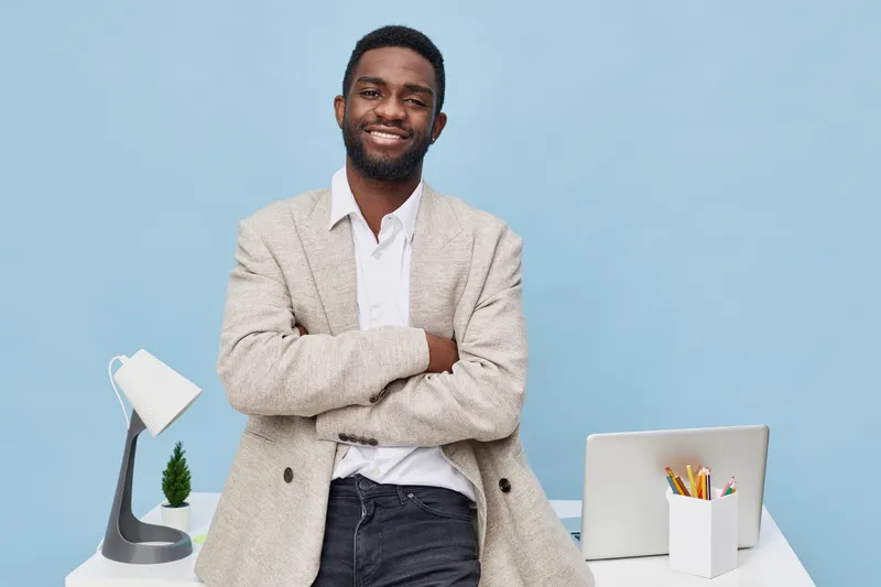 Confident smiling African American man in casual blazer and shirt standing in modern office with laptop and desk accessories, looking at camera. People lifestyle concept.