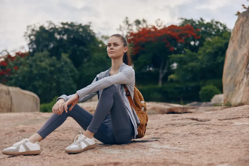 Young woman in activewear sitting on rocks, showcasing a serene expression in a nature setting with greenery and a hint of red flowers, perfect for wellness concepts