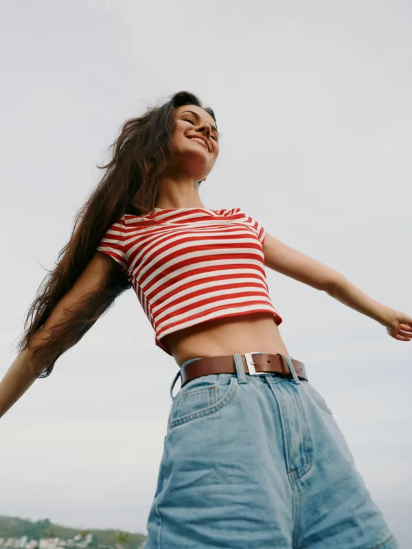 Energetic young woman spins with joy in a red striped crop top and denim jeans, wind lifting hair against a pale sky, capturing carefree youth, fashion, outdoor happiness, movement, freedom.