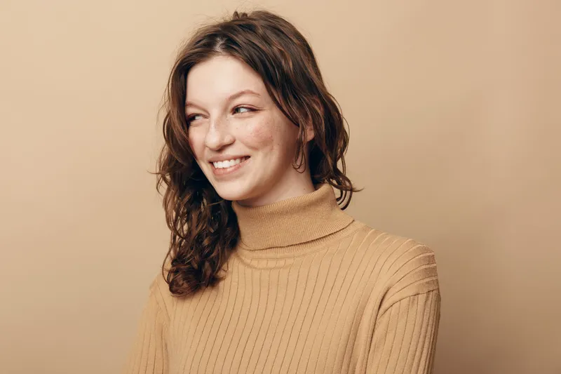 Smiling young woman with curly brown hair wearing beige turtleneck sweater looks sideways on beige background. Concept of natural beauty, casual style, and positive mood portrait.