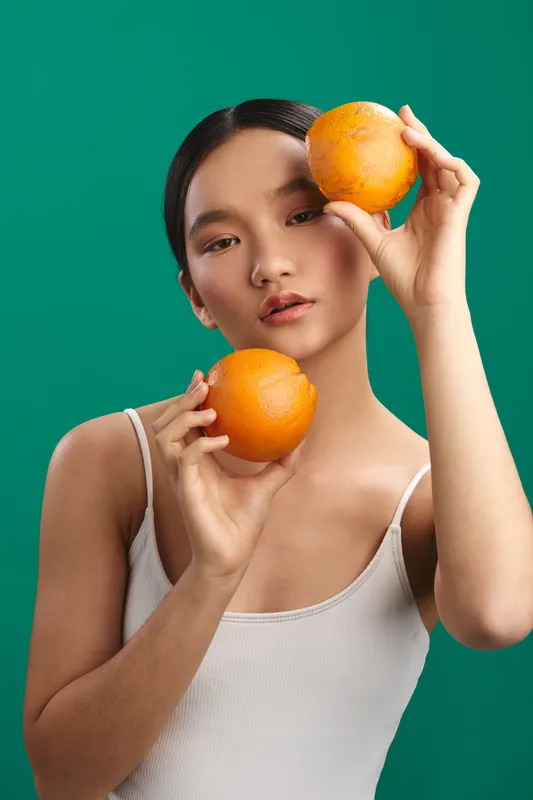 Asian woman holds bright oranges against a teal backdrop, creating a fresh still life scene. She wears a light top, eyes direct, exuding playful energy and vibrant color contrast.