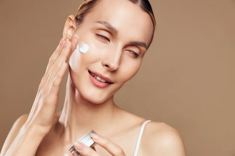 A skincare routine moment shows a woman applying moisturizer to her face, a calm ritual of hydration and selfcare in a bright, minimal setting with soft lighting.