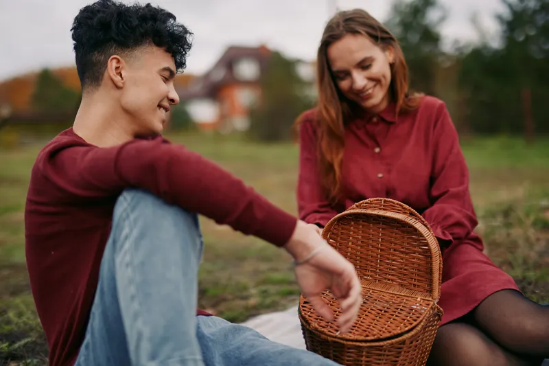A candid, authentic picnic moment of a smiling young couple sharing a woven basket outdoors, capturing genuine connection, natural interaction, believable everyday romance and simple domestic joy.