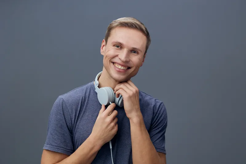 Cheerful young man holding headphones around his neck, wearing casual blue t-shirt, smiling brightly, looking at camera, isolated on plain gray background studio portrait. People lifestyle concept