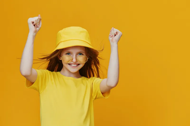 Cheerful girl celebrating with raised arms, wearing yellow clothing and accessories against a vibrant yellow background, embodying joy and positivity