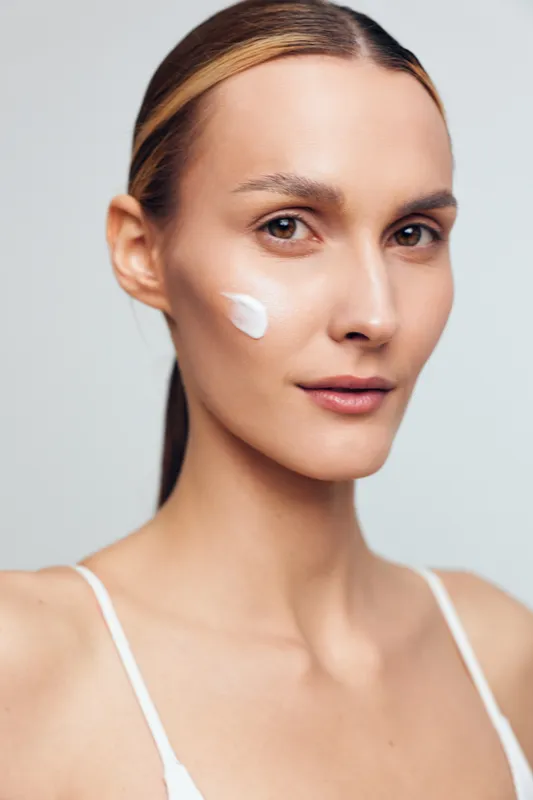 Young woman with clear skin applying white face cream on cheek. Close-up portrait with natural makeup on light background. Concept of skincare, beauty, and self-care routine.